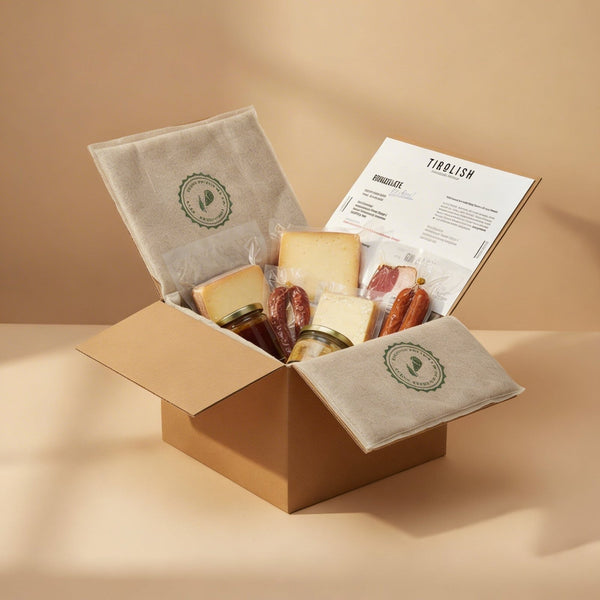 Geöffneter TIROLISH-Versandkarton mit Käsestücken, Kaminwurzen, Speck und zwei Gläsern zwischen Vlieseinlagen, dazu eine Grußkarte